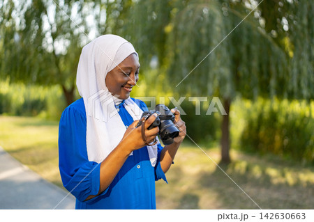 A smiling African American Muslim woman wearing a white hijab and blue dress holds a digital camera while standing in a park A smiling African American Muslim woman wearing a white hijab and blue dress holds a digital camera while standing in a park 142630663