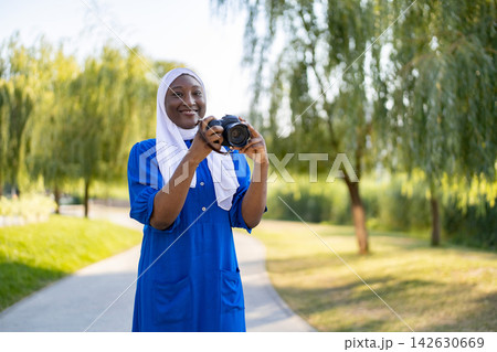 A smiling African American Muslim woman wearing a blue dress and white hijab holds a camera while standing in a park A smiling African American Muslim woman wearing a blue dress and white hijab holds a camera while standing in a park 142630669