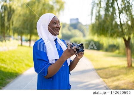 A smiling African American Muslim woman wearing a white hijab and blue dress holds a camera while standing outdoors on a sunny day A smiling African American Muslim woman wearing a white hijab and blue dress holds a camera while standing outdoors on a sunny day 142630696