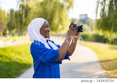 A smiling Muslim woman in a white hijab and blue dress holds a camera to capture a scenic view outdoors 142630754