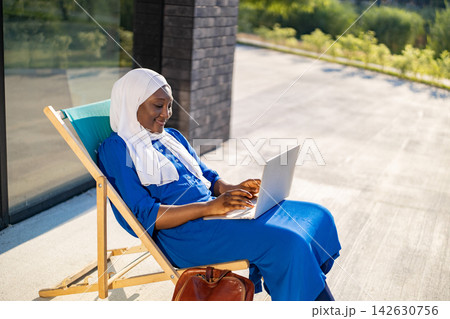 An African American Muslim woman in a hijab smiles while working on her laptop on a lounge chair outside An African American Muslim woman in a hijab smiles while working on her laptop on a lounge chair outside 142630756