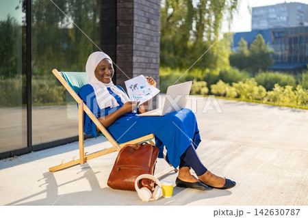 A smiling African American Muslim woman in a hijab works on her laptop while sitting in a lounge chair on a sunny day A smiling African American Muslim woman in a hijab works on her laptop while sitting in a lounge chair on a sunny day 142630787