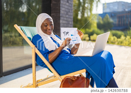A smiling African American Muslim woman in a hijab sits on a lounge chair outdoors with a laptop and documents A smiling African American Muslim woman in a hijab sits on a lounge chair outdoors with a laptop and documents 142630788