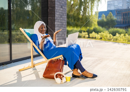 A smiling African woman wearing a hijab is engaged in a video call on her laptop while relaxing on a lounge chair outside 142630791