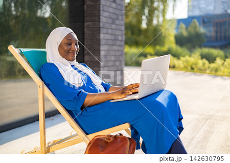 A smiling African American Muslim woman wearing a hijab and blue dress works on her laptop while relaxing on a lounge chair outside A smiling African American Muslim woman wearing a hijab and blue dress works on her laptop while relaxing on a lounge chair outside 142630795