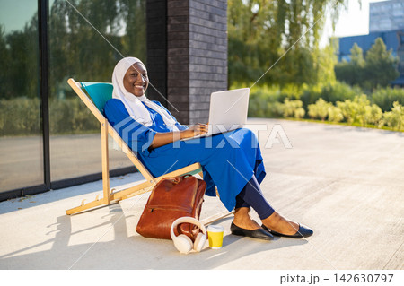 A smiling African American Muslim woman wearing a hijab and blue dress sits on a lounge chair with a laptop and backpack A smiling African American Muslim woman wearing a hijab and blue dress sits on a lounge chair with a laptop and backpack 142630797