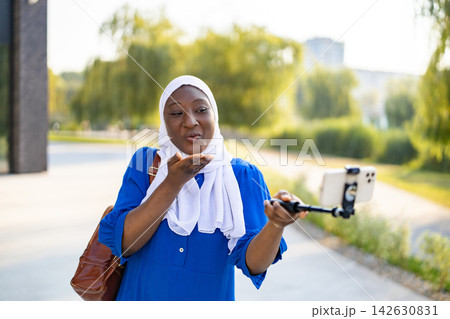 An African American Muslim woman wearing a hijab and a blue dress is taking a selfie with a smartphone on a selfie stick outdoors An African American Muslim woman wearing a hijab and a blue dress is taking a selfie with a smartphone on a selfie stick outdoors 142630831