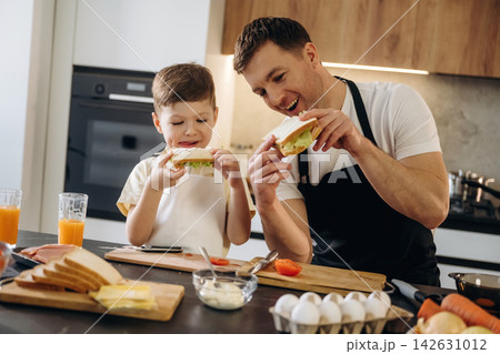 Father and son preparing sandwiches together in a modern kitchen with fresh ingredients, including bread, vegetables, and drinks on the countertop 142631012