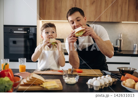 Father and son enjoying homemade sandwiches together in a modern kitchen with fresh ingredients and cooking utensils on the countertop Father and son enjoying homemade sandwiches together in a modern kitchen with fresh ingredients and cooking utensils on the countertop 142631013