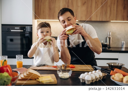 Father and son enjoying sandwiches together in a modern kitchen, surrounded by fresh vegetables, eggs, and kitchen utensils on wooden countertops 142631014