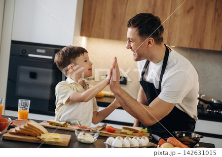 Father and young son sharing a high-five in a modern kitchen, surrounded by breakfast ingredients including bread, eggs, and vegetables on a wooden table 142631015