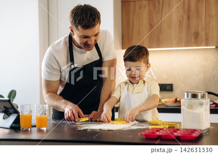 Father and son baking cookies together in a modern kitchen, using cookie cutters on a floured countertop with orange juice glasses nearby Father and son baking cookies together in a modern kitchen, using cookie cutters on a floured countertop with orange juice glasses nearby 142631330