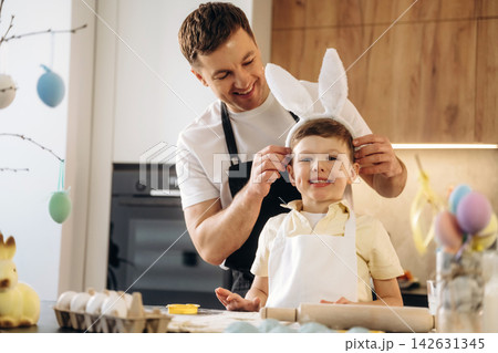 Father helps son wear bunny ears while preparing for Easter celebration in a modern kitchen with colorful decorations and eggs on the table Father helps son wear bunny ears while preparing for Easter celebration in a modern kitchen with colorful decorations and eggs on the table 142631345