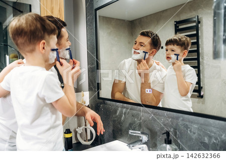 Father and son in white t-shirts shaving together in a modern bathroom, mirror reflecting their playful interaction and grooming routine Father and son in white t-shirts shaving together in a modern bathroom, mirror reflecting their playful interaction and grooming routine 142632366