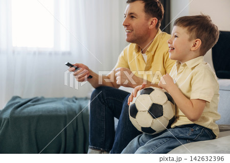 Father and son sitting on a couch watching television, smiling and holding a soccer ball, with a cozy living room setting and natural light streaming through the window 142632396