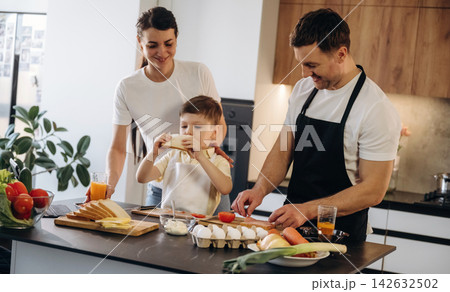 Family preparing breakfast together in a modern kitchen with fresh ingredients, including bread, eggs, and vegetables on the countertop 142632502