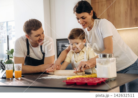 Family cooking together in a modern kitchen, mother and father assisting young boy with rolling dough on countertop surrounded by ingredients and kitchenware 142632511