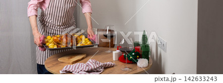 A man prepares a pre-Christmas dinner in his kitchen. He places a tray of baked chicken and potatoes on the table. Surrounding decor hints at a festive atmosphere, banner A man prepares a pre-Christmas dinner in his kitchen. He places a tray of baked chicken and potatoes on the table. Surrounding decor hints at a festive atmosphere, banner 142633243