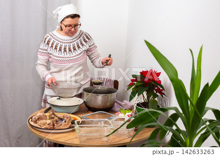 A woman stands in her kitchen preparing kholodets, aspic, a traditional Ukrainian dish. She strains the broth from the bones into a bowl while wearing comfortable clothes A woman stands in her kitchen preparing kholodets, aspic, a traditional Ukrainian dish. She strains the broth from the bones into a bowl while wearing comfortable clothes 142633263