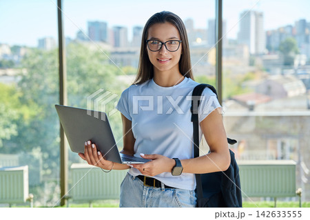 Smiling female student wearing eyeglasses holding laptop with city window background 142633555