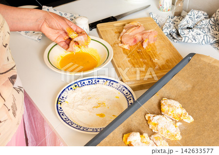 In a kitchen, a woman dips chicken breast pieces in egg while preparing to make deep-fried chicken nuggets. Flour sits on a plate beside her In a kitchen, a woman dips chicken breast pieces in egg while preparing to make deep-fried chicken nuggets. Flour sits on a plate beside her 142633577