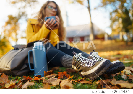 Tourist sits in clearing among yellow fallen leaves, drinks tea from thermos in autumn park. Tourist sits in clearing among yellow fallen leaves, drinks tea from thermos in autumn park. 142634484