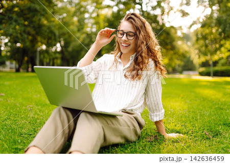 Young woman in glasses and with laptop working at sunset in park on green meadow. Lifestyle concept. Young woman in glasses and with laptop working at sunset in park on green meadow. Lifestyle concept. 142636459