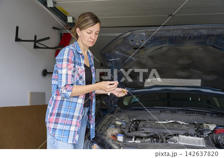 Woman examining engine oil dipstick beside car with open hood in home garage Woman examining engine oil dipstick beside car with open hood in home garage 142637820