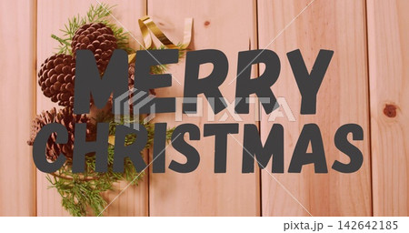 Dominating dark-gray MERRY CHRISTMAS text over pine planks, pine cones, fir sprigs, gold ribbon Dominating dark-gray MERRY CHRISTMAS text over pine planks, pine cones, fir sprigs, gold ribbon 142642185