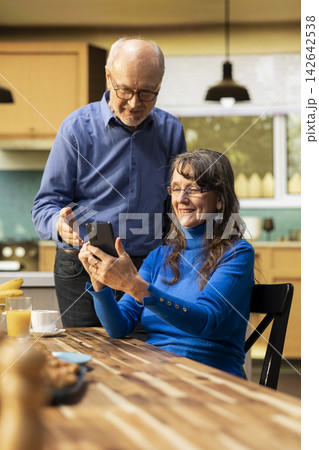 Married people scrolling apps on smartphone and sitting together in the kitchen, enjoying fresh coffee drink and casual conversation indoors. Casual moment during a relaxed family morning. 142642538