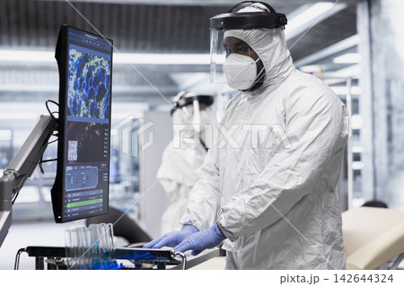 Specialist in quarantine performing clinical study on an infectious patient, testing vaccine responses and monitoring DNA molecules on monitor to develop treatments for epidemics. 142644324
