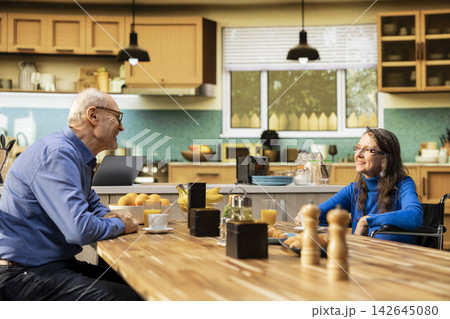 Old woman with disability and husband serve coffee and chatting at kitchen table, candid moment reflecting healthy lifestyle and comfort in retirement age. Elderly pair share conversation. Old woman with disability and husband serve coffee and chatting at kitchen table, candid moment reflecting healthy lifestyle and comfort in retirement age. Elderly pair share conversation. 142645080