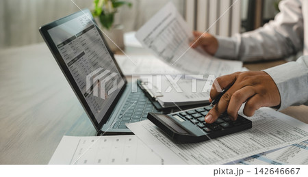 businessmen using calculator while holding pen over printed financial reports beside laptop, showing financial calculation, report review, data analysis and accounting workflow. businessmen using calculator while holding pen over printed financial reports beside laptop, showing financial calculation, report review, data analysis and accounting workflow. 142646667