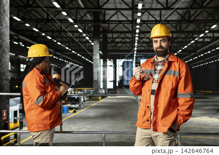 Engineer speaks into portable walkie-talkie inside heavy industry warehouse, wearing hard hat. Wireless radio device enables clear communication and coordination across large-scale machinery hall. Engineer speaks into portable walkie-talkie inside heavy industry warehouse, wearing hard hat. Wireless radio device enables clear communication and coordination across large-scale machinery hall. 142646764