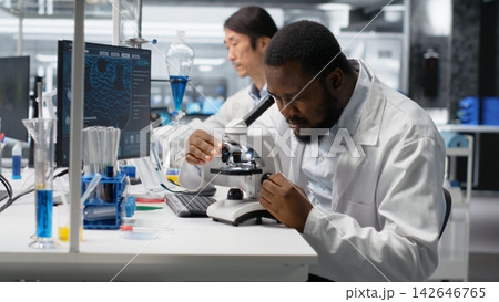 Researcher operating compound microscope, doing sample observation in controlled laboratory environment. African american man studies tissue under optical device in biotechnology lab, camera B Researcher operating compound microscope, doing sample observation in controlled laboratory environment. African american man studies tissue under optical device in biotechnology lab, camera B 142646765