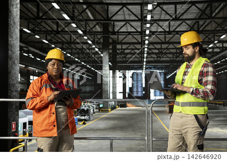 Engineering plant staff with hard hat inspecting heavy machinery and production line on laptop, industrial factory. Massive warehouse facility with steel beams and metallic framework. Engineering plant staff with hard hat inspecting heavy machinery and production line on laptop, industrial factory. Massive warehouse facility with steel beams and metallic framework. 142646920