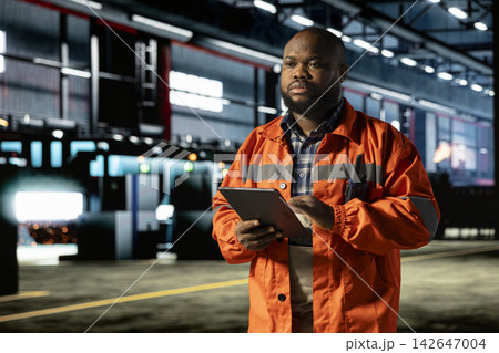 African american man manages heavy machinery activity on tablet in manufacturing facility, wearing protective gear and walking in the factory. Labor strength for engineering operations. 142647004