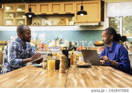 Young black couple planning grocery orders online using a laptop. Boyfriend and girlfriend discuss shopping list and comparing supermarket prices, choosing items for consumption needs. 142647068