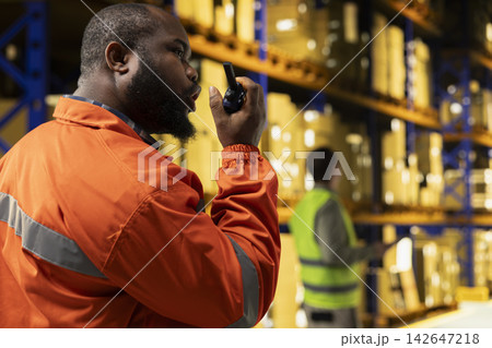 Black warehouse operator using portable walkie-talkie radio in distribution hub. Staff coordinates shipment details, parcel tracking and cargo handling with two-way transmitter in depot. Black warehouse operator using portable walkie-talkie radio in distribution hub. Staff coordinates shipment details, parcel tracking and cargo handling with two-way transmitter in depot. 142647218