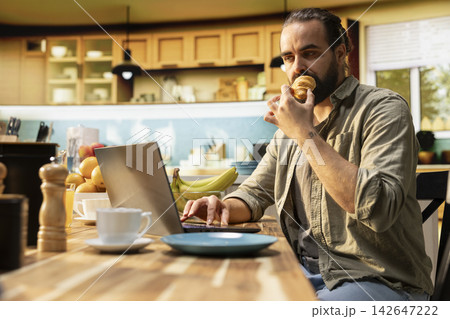 Remote worker, eating croissants, using laptop in kitchen, solving daily tasks online, serving breakfast. Digital nomad, freelancer lifestyle with flexible work, multitasking and teleworking. 142647222