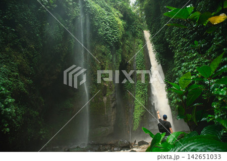 A traveler admires a tall waterfall in lush greenery 142651033