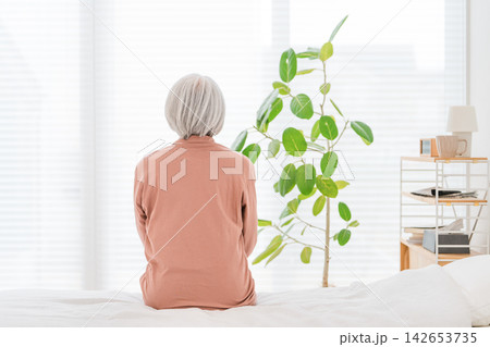 ベッドに座り窓の外を見つめるシニア女性。老後の孤独や将来への不安を抱える高齢者の後ろ姿 142653735