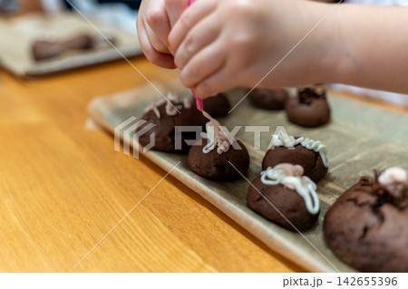 子供が白と薄茶色のアイシングでチョコクッキーをデコレーションしている手元の写真 子供が白と薄茶色のアイシングでチョコクッキーをデコレーションしている手元の写真 142655396