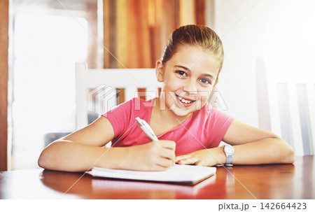 Portrait, happy child and writing at table for school, studying and education in house for knowledge. Notebook, girl and student for learning, exam and homeschool for development with pride 142664423