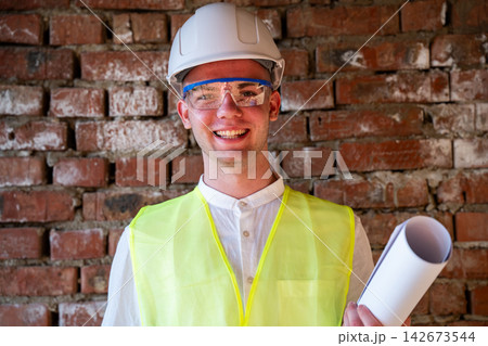 Smiling construction professional wearing hard hat and safety vest while holding rolled plans against a brick wall. Architecture renovation planning and building supervision 142673544