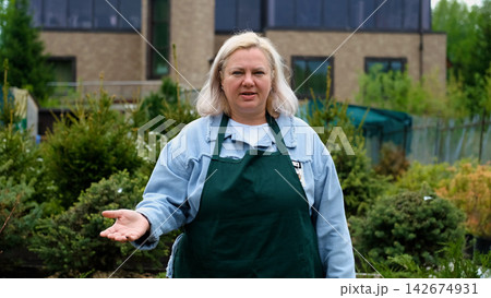 Portrait of happy female gardener standing among bright greenery of her garden Portrait of happy female gardener standing among bright greenery of her garden 142674931