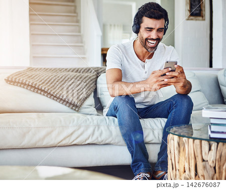 Man, headphones and laughing with phone in home for funny video, social media post and comedy movie on mobile app. Person, smartphone and happy for listening to audio, sound and subscription on couch 142676087