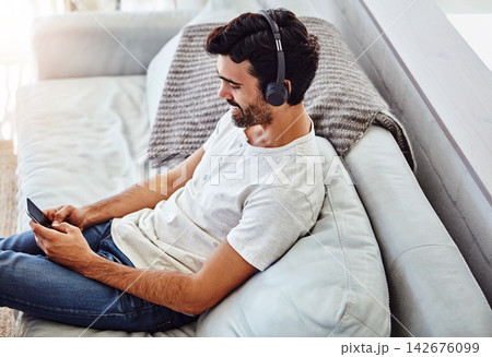 Home, man and cellphone with headphones on couch for listening to music, radio and networking. Living room, male chatter and mobile app for streaming podcast, online games and social media to relax 142676099