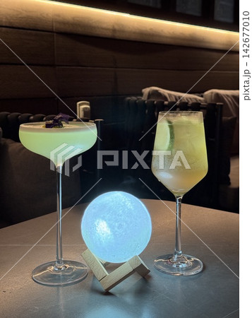 Fancy colorful cocktails and a glowing orb moon lamp on a wooden table in a cozy dimly lit bar lounge 142677010