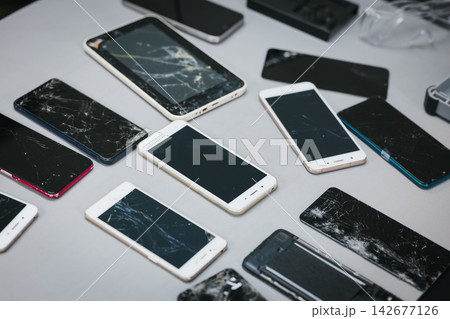 Collection of broken smart phones with cracked screens scattered on a table, device damage, repair needs, and electronic waste. 142677126